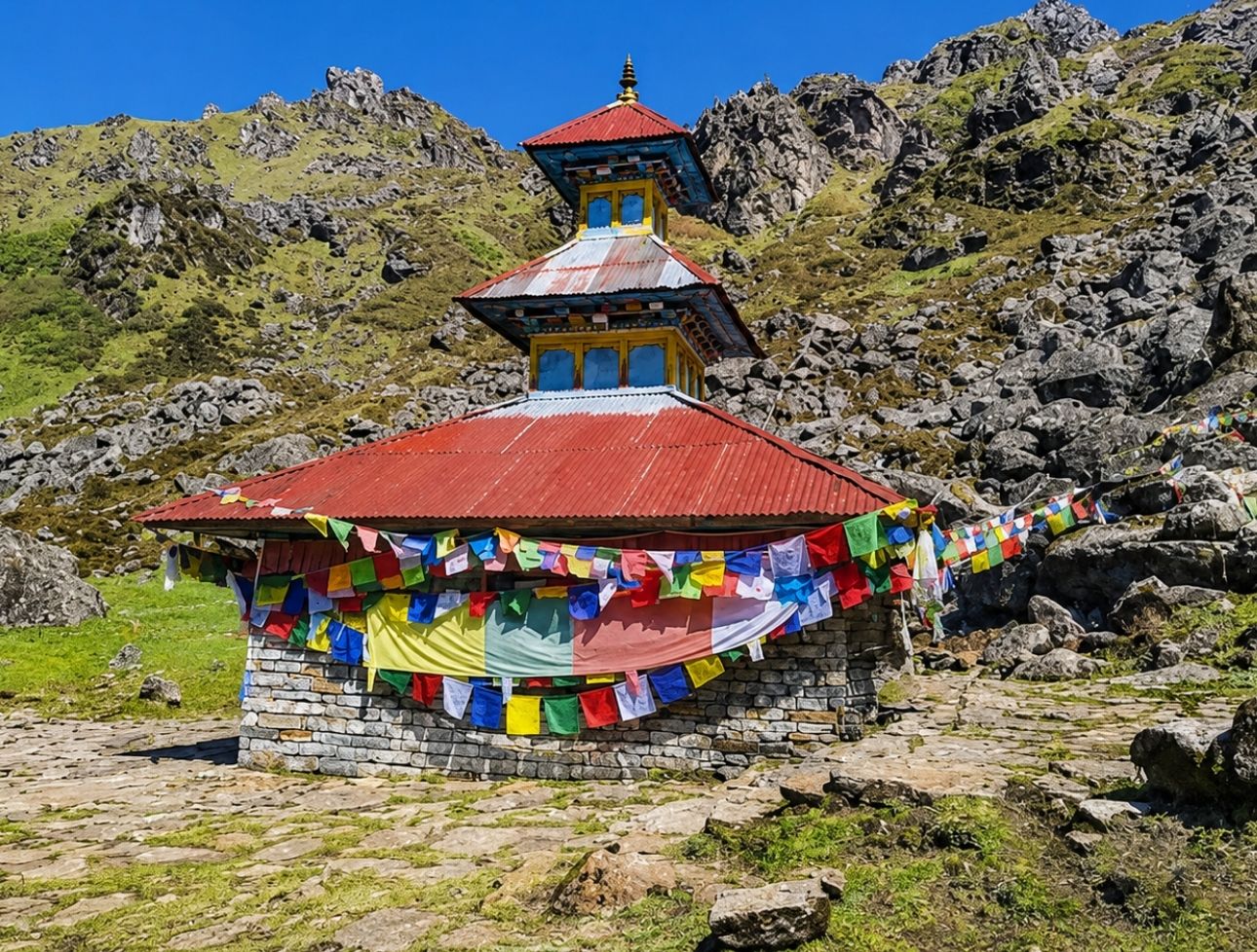 A traditional Pagoda-style Hindu temple made of stone and a red metal roof, adorned with colorful Buddhist prayer flags, set against a rocky mountain slope in Panchpokhari, Nepal.