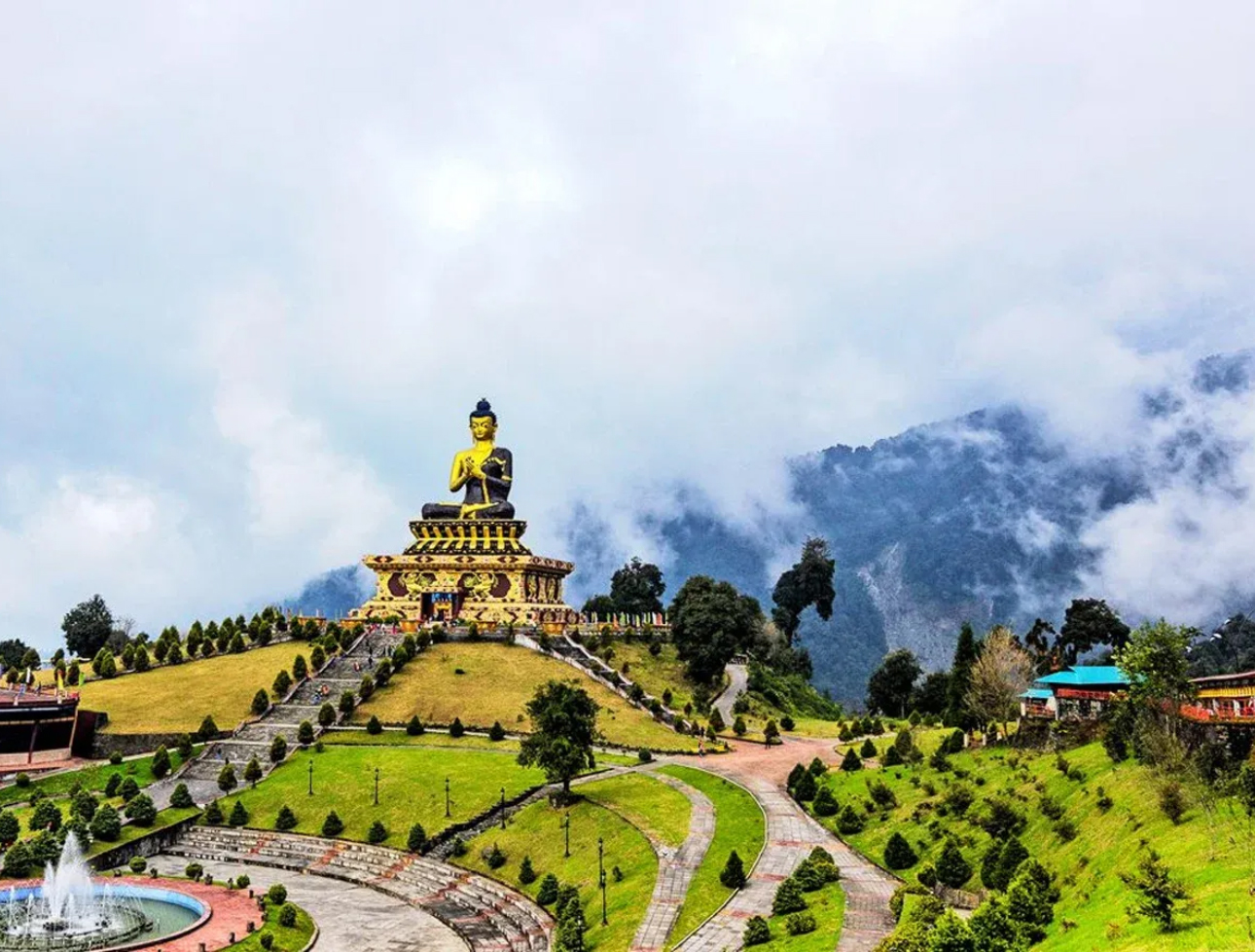 A large Buddha statue standing prominently on a grassy hill with cloudy mountains in the background in Kalimpong.