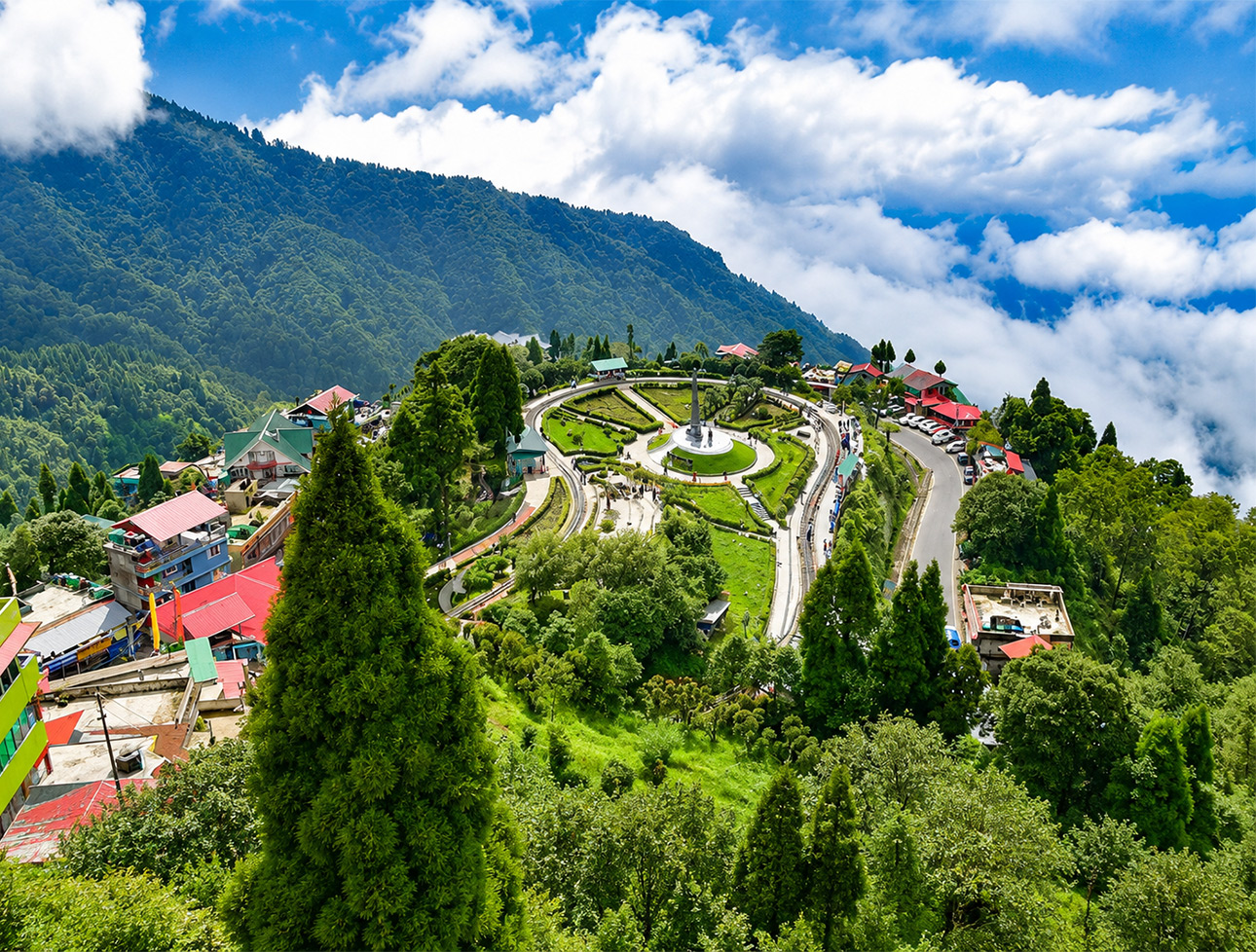 A winding road leading through a hillside town in Darjeeling, surrounded by lush green trees and distant mountains under a blue sky.